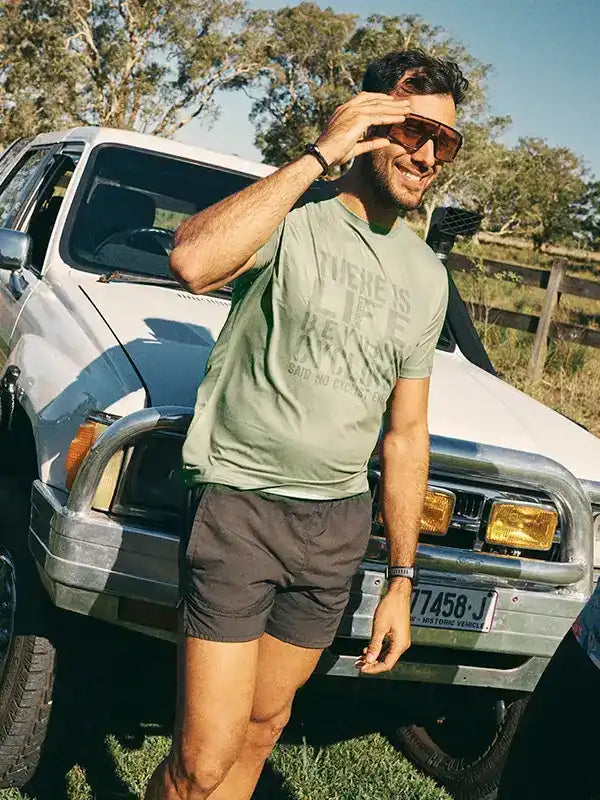 Man in olive cycling tee and shorts leaning on white truck outdoors, stylish men's bike apparel