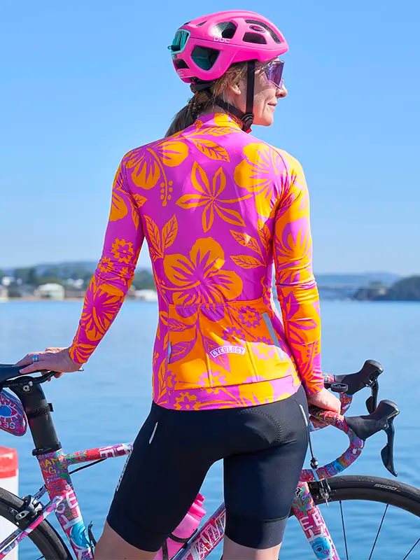 Person wearing Oahu Pink Women's Long Sleeve Summer Cycling Jersey standing next to a bicycle by a body of water.