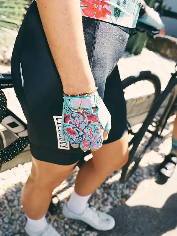 Cyclist wearing floral-patterned cycling gloves and black shorts, standing beside bike outdoors