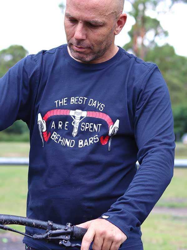 Man wearing navy long-sleeve cycling shirt with 'The Best Days Are Spent Behind Bars' graphic, holding bike handlebars outdoors