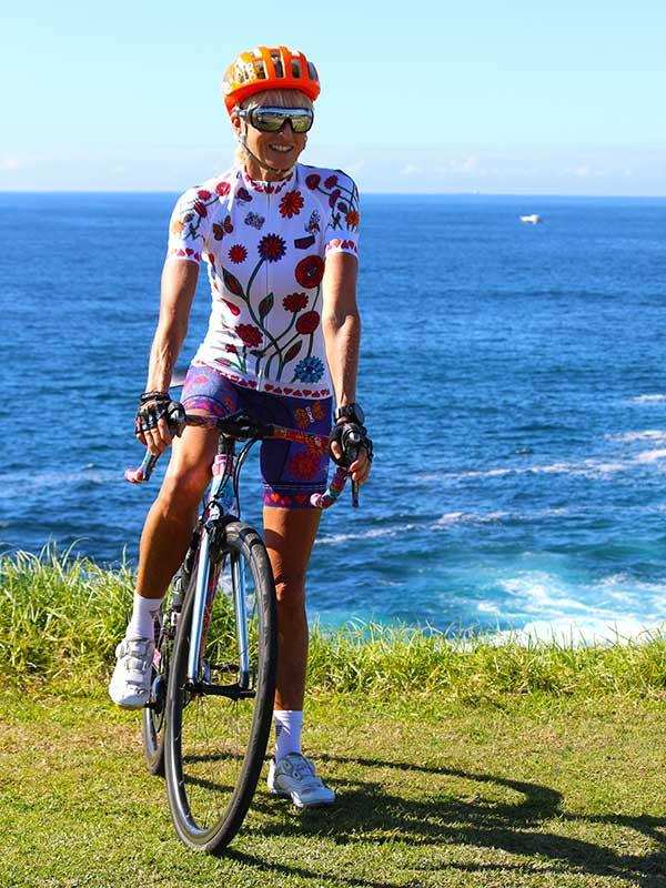 Woman in floral cycling jersey and shorts riding bike by ocean, wearing orange helmet and sunglasses