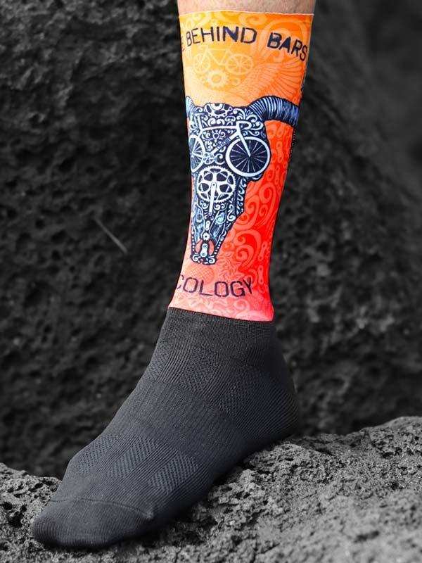 Cycling sock with bull skull & bike design, orange gradient, 'Behind Bars' text, worn on foot outdoors