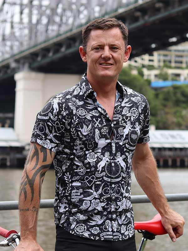 Man wearing black and white cycling-themed shirt, holding red bike near bridge