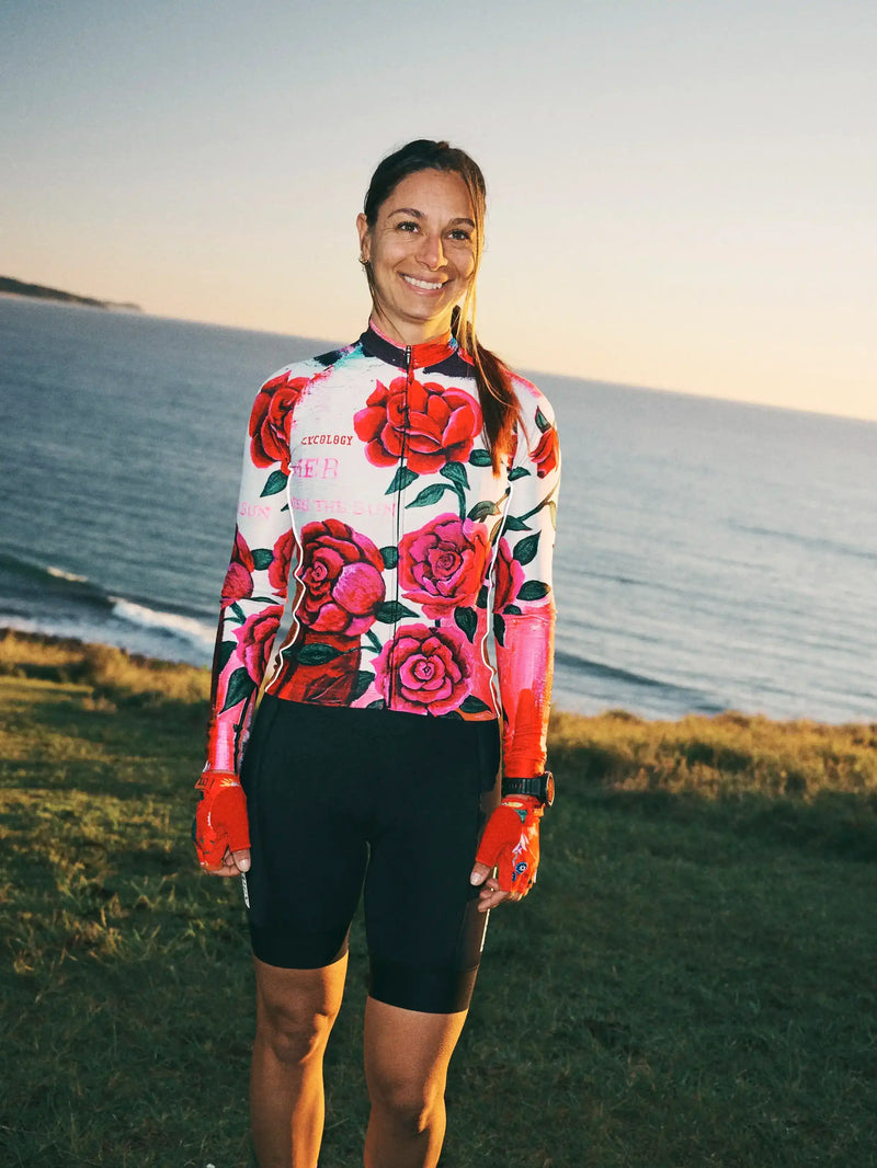 Woman in floral long sleeve cycling jersey and black shorts, standing by the sea at sunset.