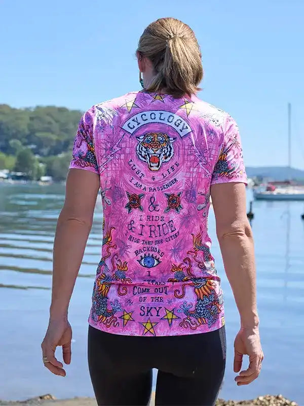 Woman wearing vibrant pink cycling jersey with tiger graphic and 'I Ride' text, standing by water