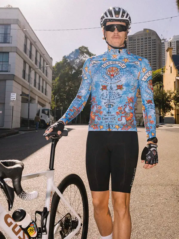Cyclist in a colorful long sleeve blue Summer jersey and black shorts standing next to a bicycle on a city street.