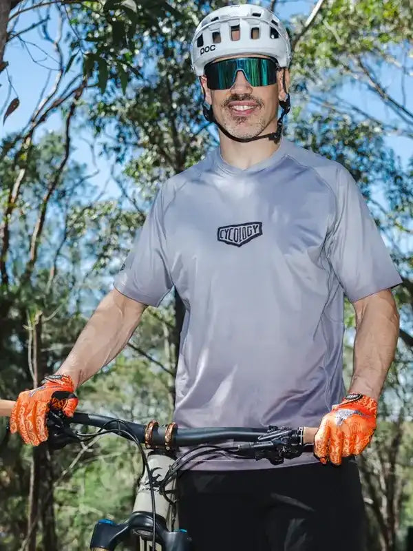 Man in grey cycling jersey and orange gloves on mountain bike trail, wearing POC helmet and sunglasses
