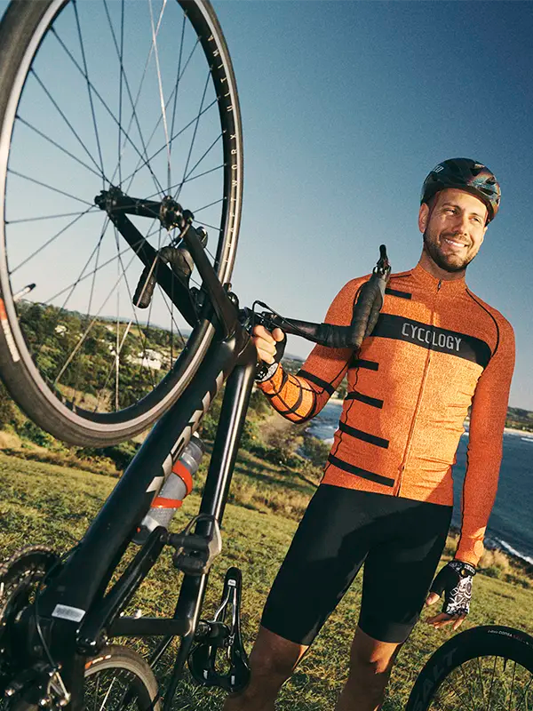 Cyclist in orange and black cycling outfit holding a bicycle outdoors with a scenic background.