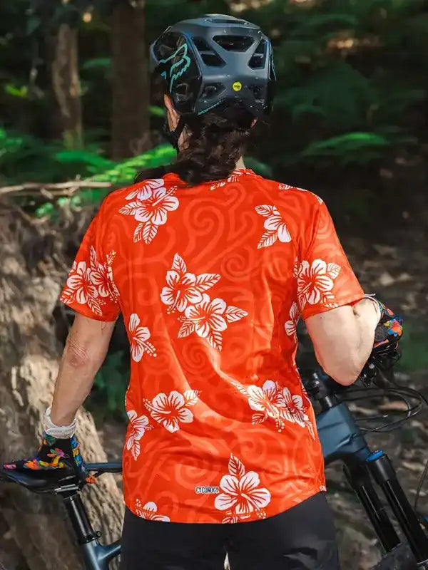Woman in orange floral cycling jersey riding mountain bike in forest, wearing helmet and gloves