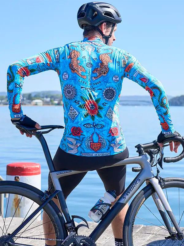 Person wearing Koa Samui Men's Blue Long Sleeve Summer Cycling Jersey a colorful cycling jersey and helmet with a bicycle by a waterfront.
