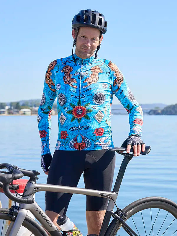 Man in Koa Samui Men's Blue Long Sleeve Summer Cycling Jersey standing next to a bicycle by a body of water.