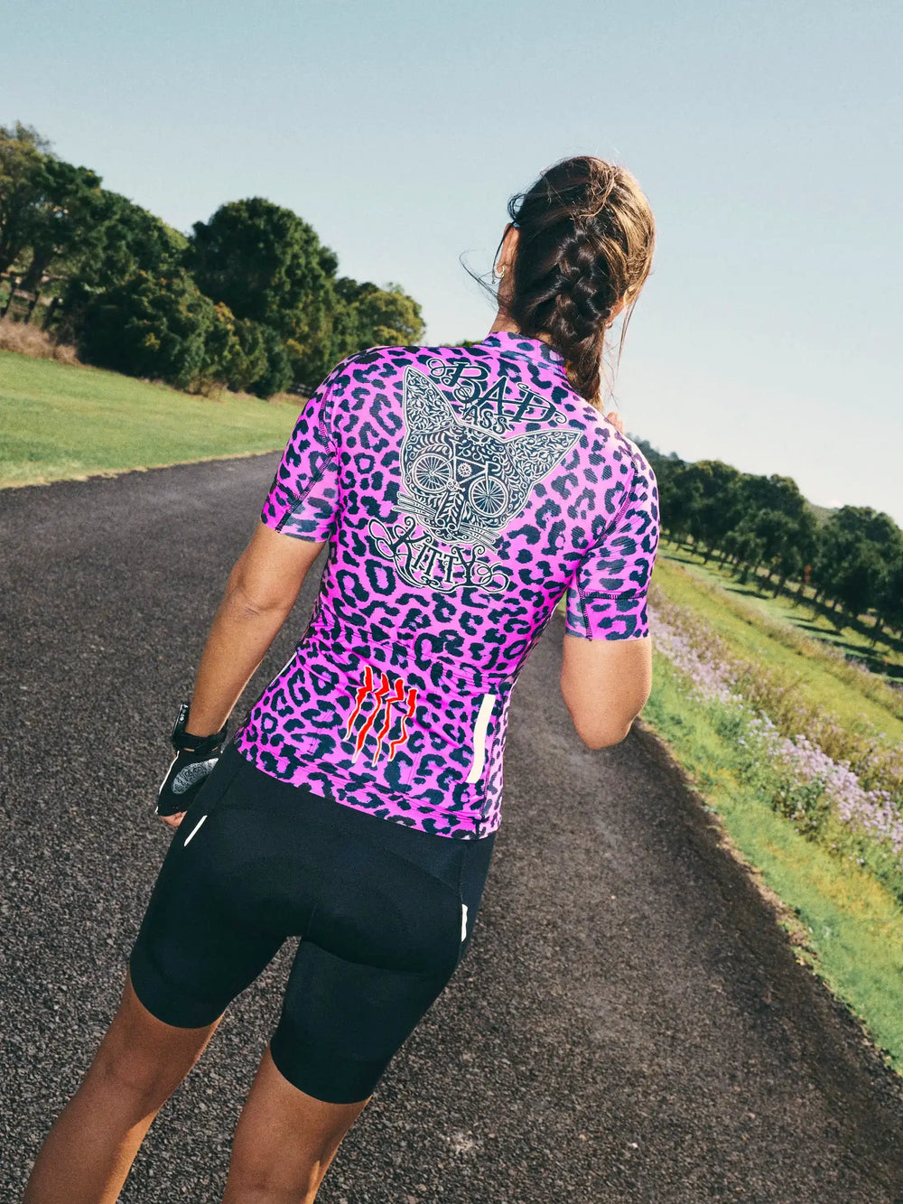 Woman cycling in vibrant pink leopard print jersey with 'Badass Kitty' graphic, black shorts, on scenic road.