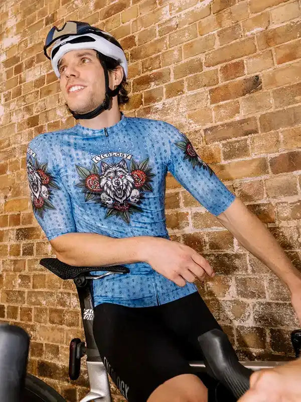Man wearing blue cycling jersey with wolf and rose design, black shorts, helmet, on bike against brick wall