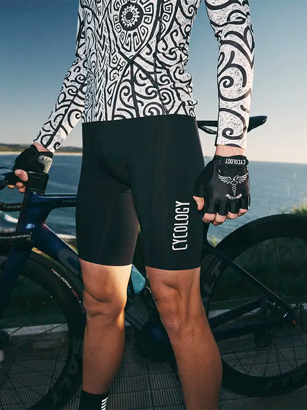 Person wearing black cycling bib shorts with 'Cycology' branding, standing next to a bicycle by a body of water.