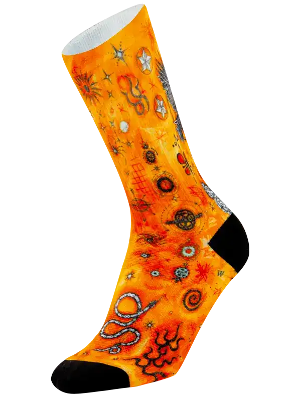 A single crew cycling sock with a vibrant orange and yellow background featuring a detailed Aboriginal-style art pattern of snakes, stars, and geometric symbols, finished with solid black toes and heels.