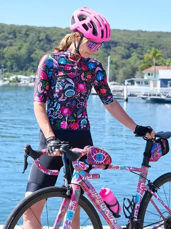Woman in vibrant floral cycling jersey and pink helmet, standing with matching bike by waterfront