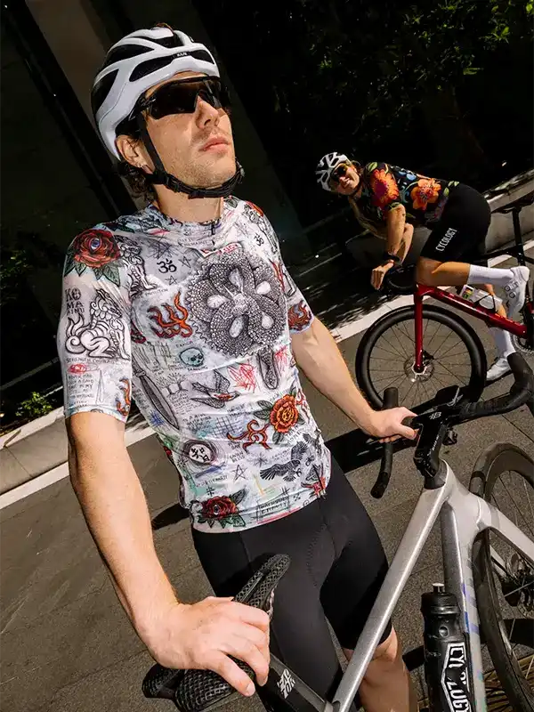 Man in tattoo-style cycling jersey and helmet, riding road bike with woman in floral kit behind