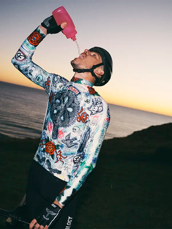 Person wearing a colorful long sleeve jersey  with a helmet, pouring a pink bottle against a sunset sky.