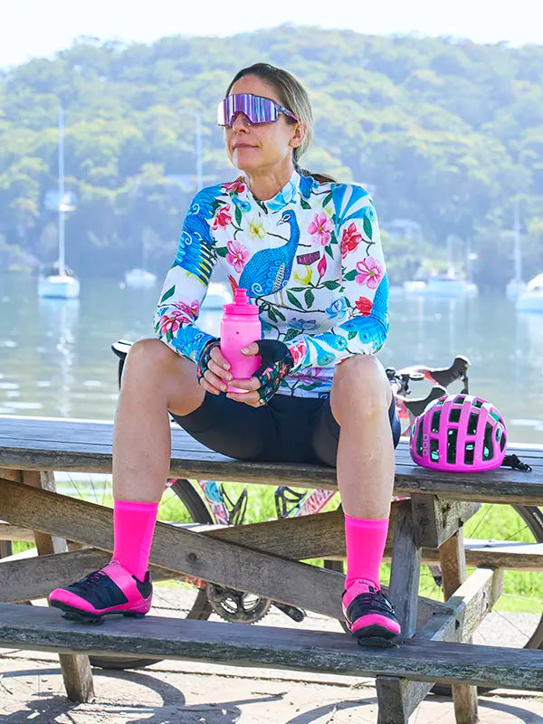 Woman sitting on a picnic table with a scenic background, wearing Goddess White Women's Long Sleeve Summer Cycling Jersey with peacock and floral pattern on a white background