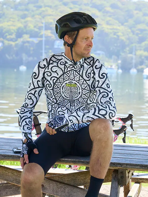 Man sitting on a bench wearing a patterned cycling jersey and helmet with a scenic background.