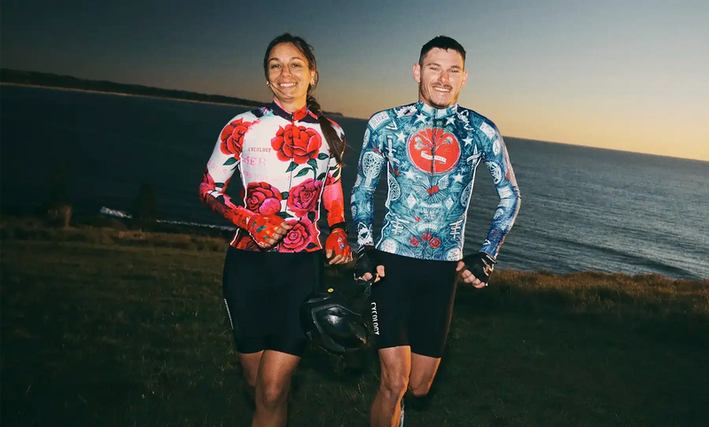 Man and woman in vibrant cycling jerseys and shorts, standing by the sea at sunset, holding helmets.