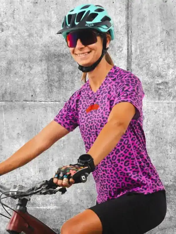 Woman cycling in vibrant pink leopard print jersey, helmet, and sunglasses on urban concrete background