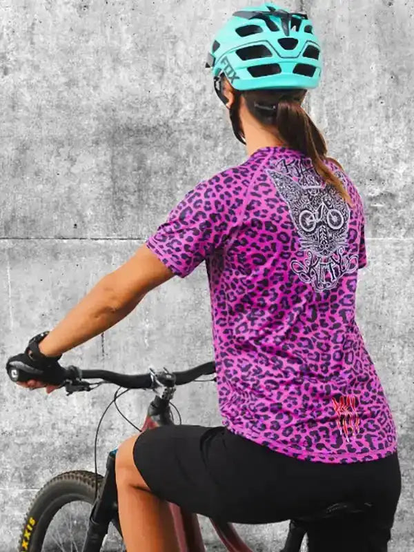 Woman cycling in vibrant pink leopard print jersey with owl graphic, wearing teal helmet and black shorts