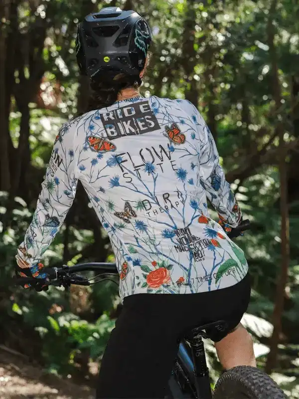 Woman riding mountain bike in floral long sleeve cycling jersey with 'RIDE BIKES' print, forest trail backdrop