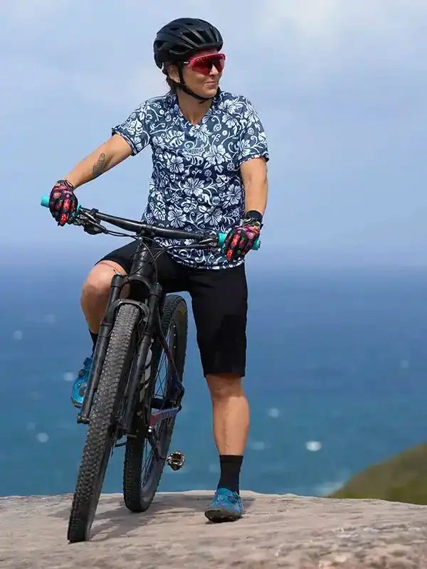 Man in floral cycling jersey and shorts riding mountain bike by ocean, wearing helmet and gloves