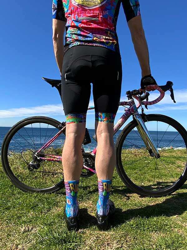 Cyclist in colorful cycling jersey and matching socks standing with road bike by ocean, grassy cliffside