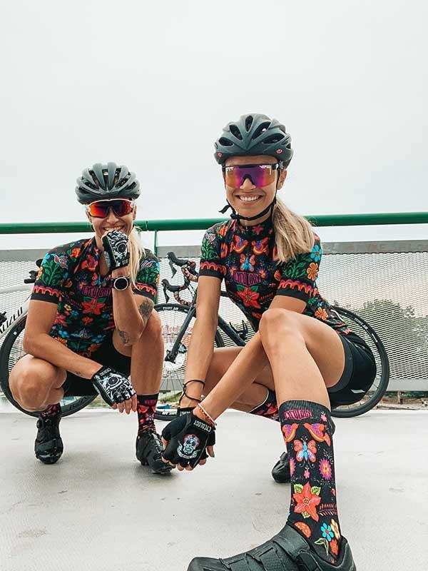 Women in colorful floral cycling jerseys, socks, and helmets posing with bikes outdoors