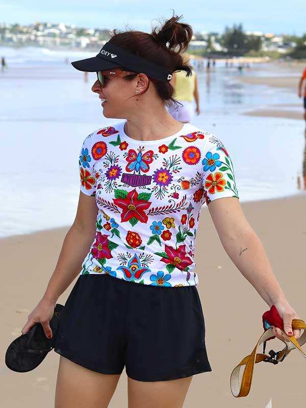 Woman in colorful floral cycling jersey and black shorts walking on beach, holding sandals