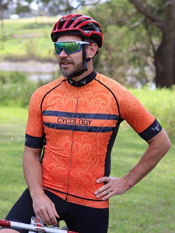 Man in orange patterned cycling jersey with 'CYCOLOGY' logo, wearing helmet and sunglasses outdoors