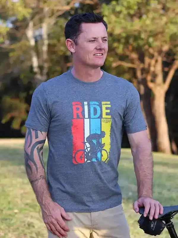 Man wearing gray cycling t-shirt with 'RIDE' graphic and cyclist design, standing outdoors near bike