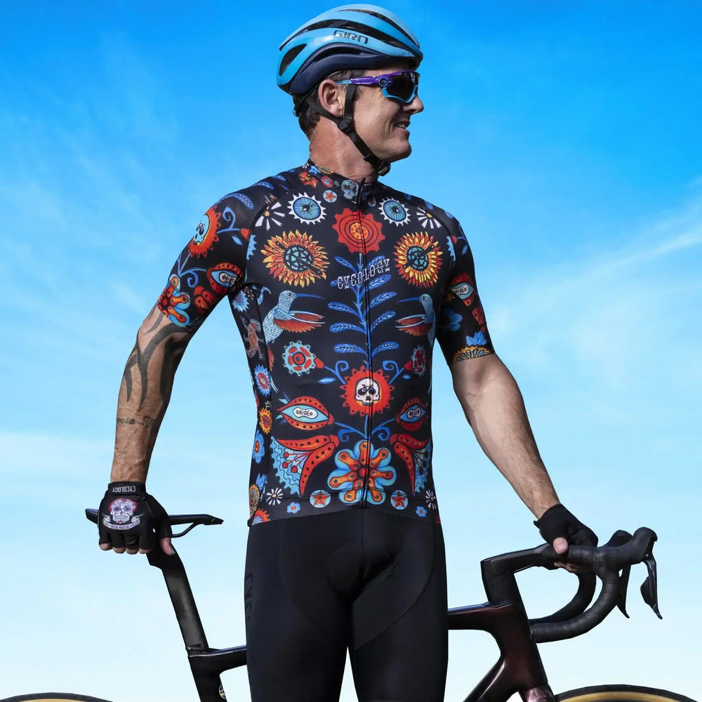 Man in vibrant floral cycling jersey and helmet, holding road bike against blue sky