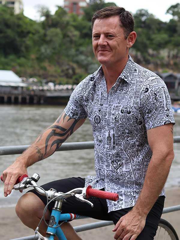 Man wearing graphic cycling shirt on blue bike by waterfront, stylish men's cycling apparel